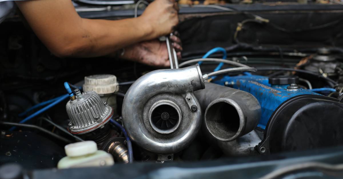 A worker moves a wrench under the hood of a truck with a silver turbocharger standing out from all other components.